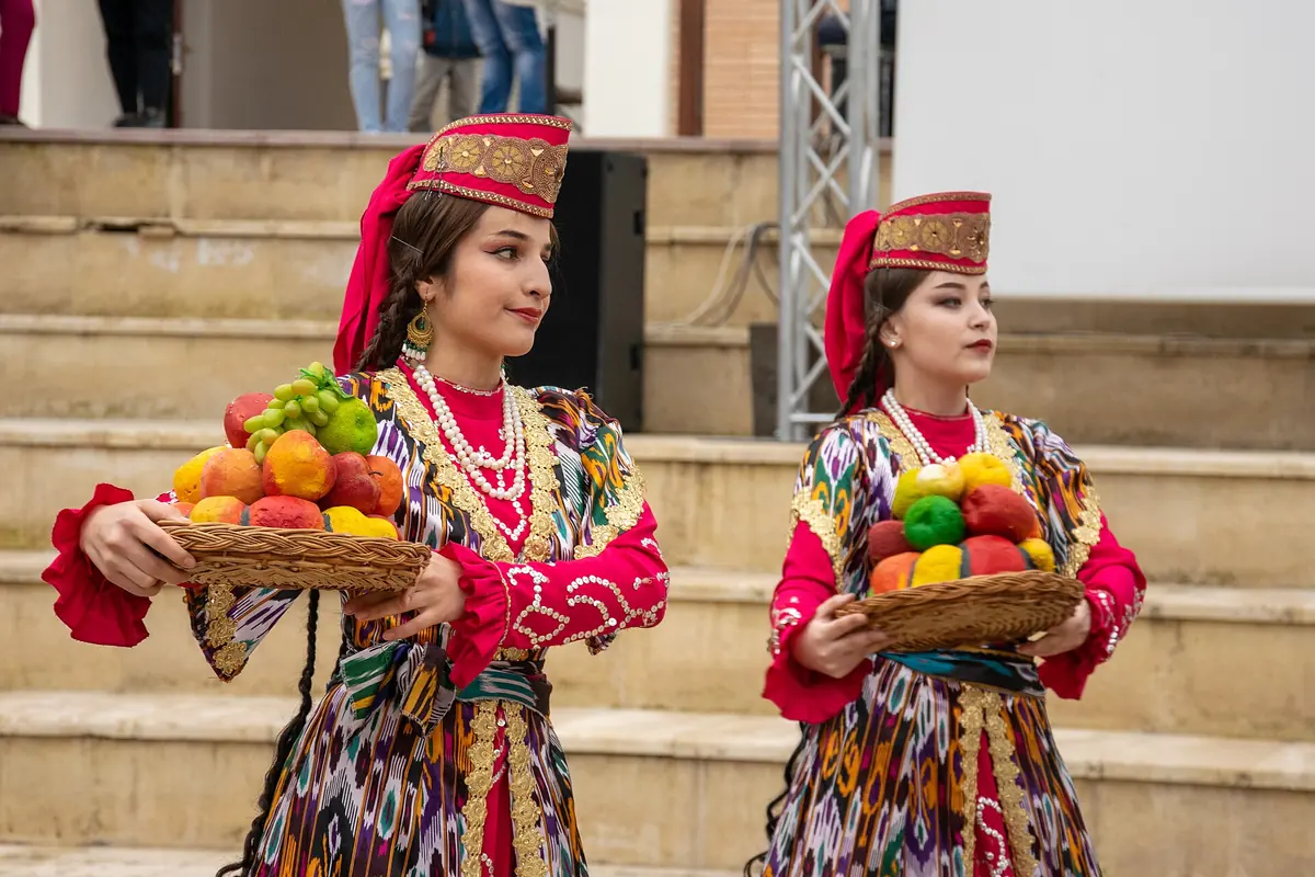 Two dancers in ikat dresses with gold embroidery, red caps and long braids hold baskets of fruit on stone steps